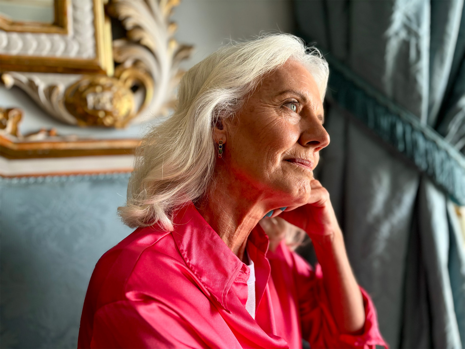 Mode portrait iPhone 15 Femme mûre, les cheveux blancs arrivant à ses épaules et vêtue d'un chemisier rose, regardant par la fenêtre, se tenant la tête avec la main sous le menton.