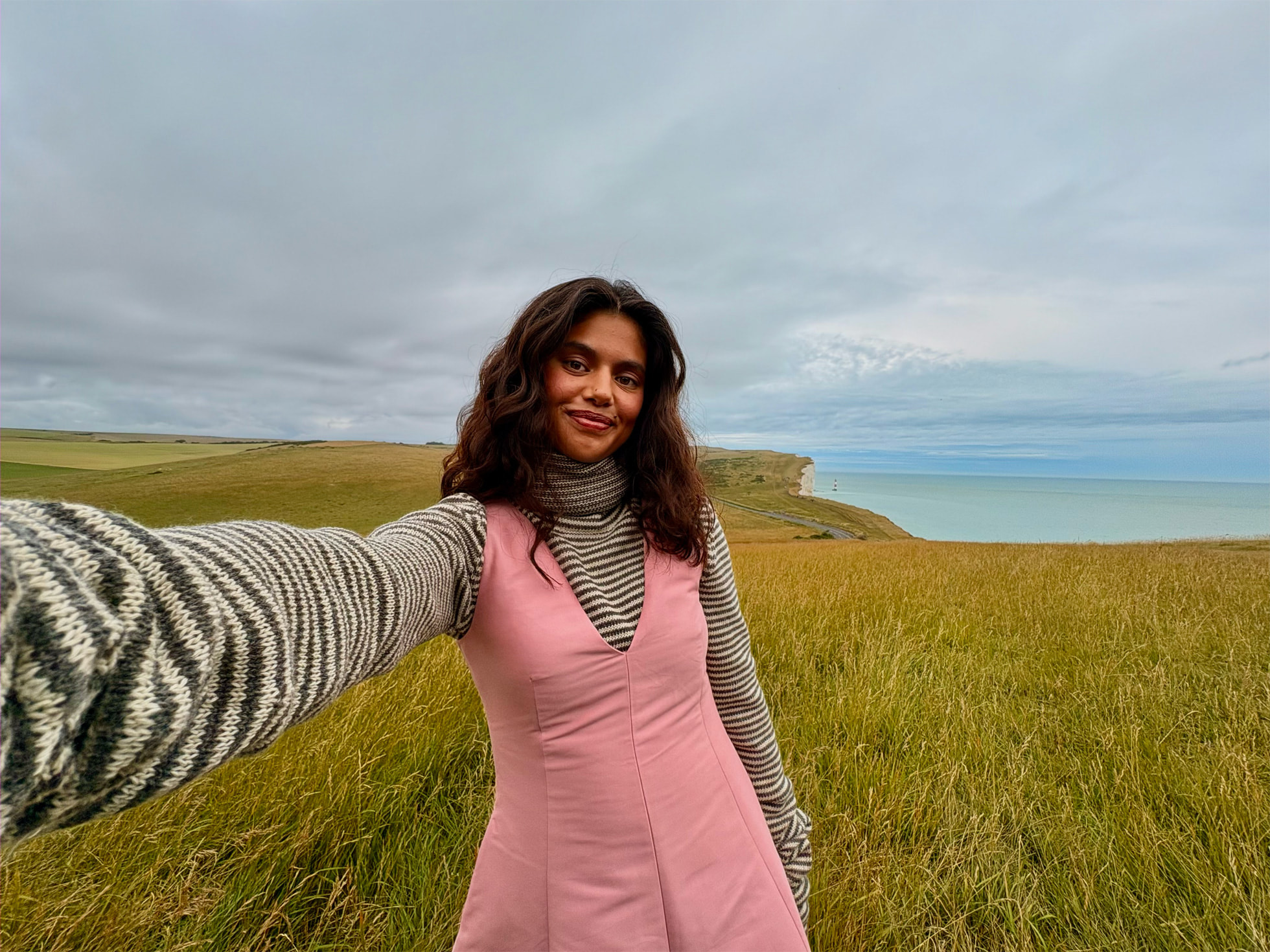 Caméra ultra grand-angle iPhone 16 Plus - Macavenue Saumur Un selfie d'une femme en robe rose avec un sous t-shirt zébré avec en arrière-plan un paysage verdoyant menant à la mer.